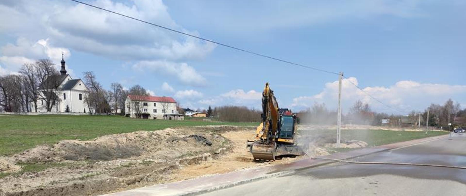 Obok uszkodzonego rurociągu ustawiona jest kurtyna wodna przez którą przepływa woda tworząc parasol ochronny. Obok zaistniałego zdarzenia znajduje się koparka.