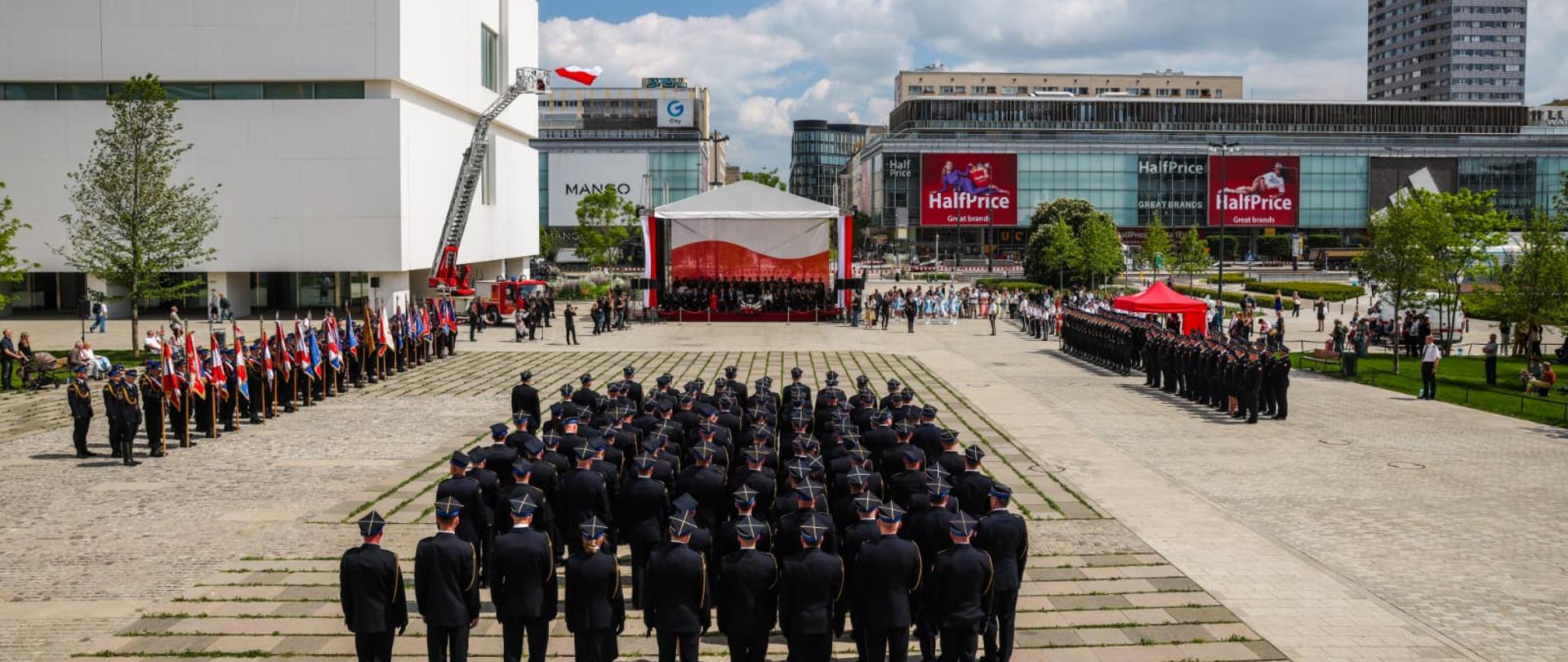 Na zdjęciu widać strażaków ustawionych w zwartym szyku na Placu Centralnym w Warszawie. W tle znajdują się nowoczesne budynki oraz scena przygotowana do uroczystości. Strażacy mają na sobie mundury wyjściowe i stoją naprzeciw trybuny honorowej.
