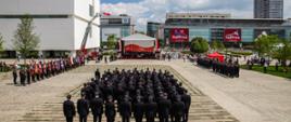 Na zdjęciu widać strażaków ustawionych w zwartym szyku na Placu Centralnym w Warszawie. W tle znajdują się nowoczesne budynki oraz scena przygotowana do uroczystości. Strażacy mają na sobie mundury wyjściowe i stoją naprzeciw trybuny honorowej.