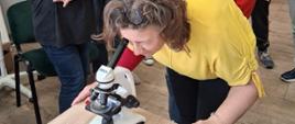 Grupa uczestników zajęć edukacyjnych zgromadzonych wokół stołu, na którym znajduje się mikroskop oraz materiały dydaktyczne. Jedna z osób pochyla się nad mikroskopem, prowadząc obserwację preparatu. Wokół stołu stoi kilka innych osób, uważnie obserwujących przebieg ćwiczenia. Na ich ubraniach widoczne są imienne identyfikatory. W tle znajdują się przeszklone gabloty z różnymi eksponatami, dekoracjami i przedmiotami z pracowni, a także biała tablica ustawiona na sztaludze.
Scena ukazuje praktyczną część zajęć z profilaktyki zdrowotnej, podczas której uczestnicy mogą samodzielnie korzystać z mikroskopu i zapoznawać się z preparatami edukacyjnymi.