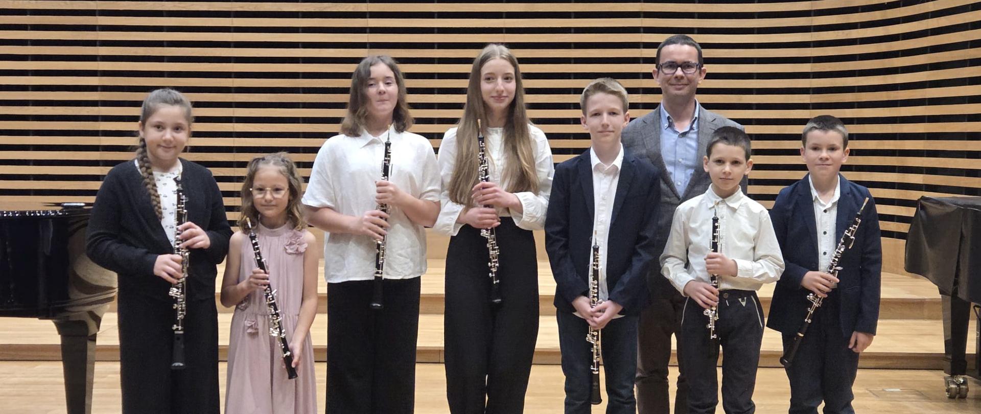 Uczniowie stoją z nauczycielem na scenie i trzymają w rękach swoje instrumenty.