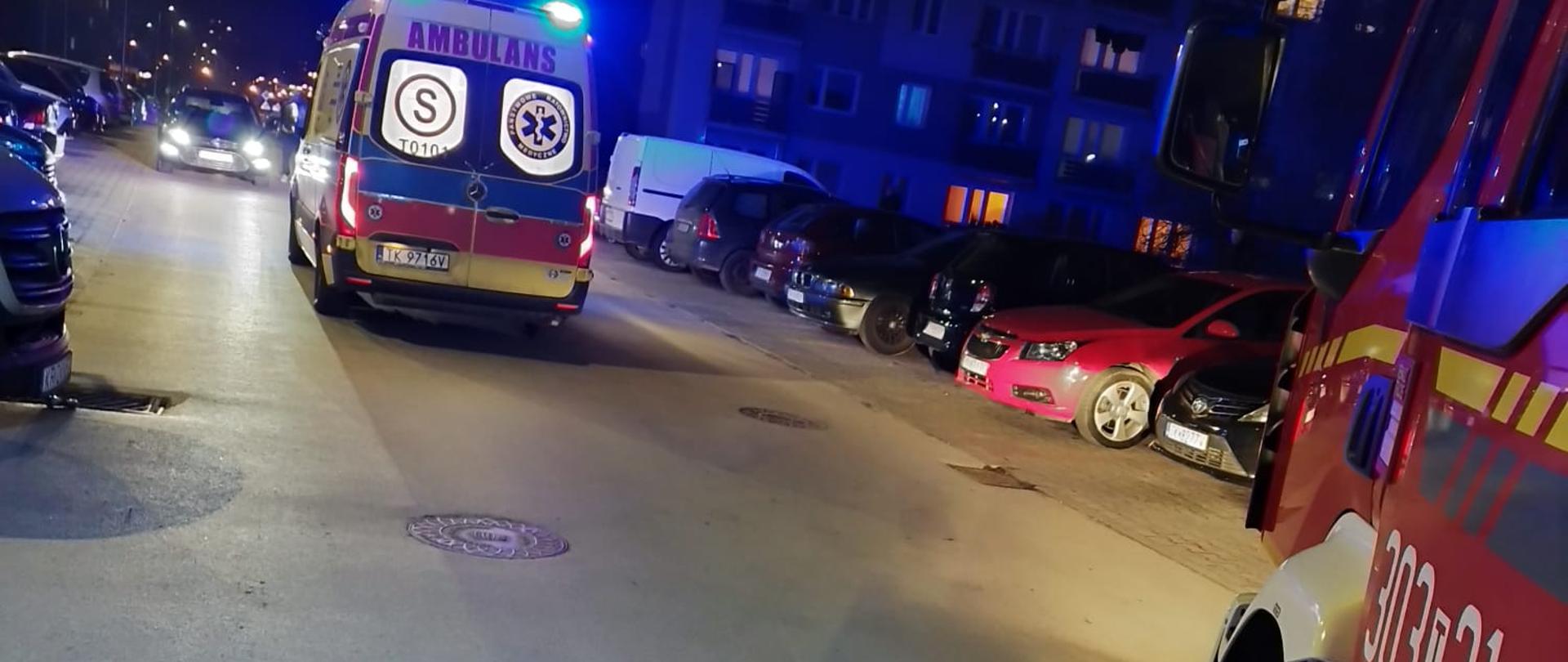 Zdjęcie przedstawia ulicę Szkolną. W centralnym punkcie fotografii widać ambulans Państwowego Ratownictwa Medycznego, a po prawej pokazano fragment wozu strażackiego. Wzdłuż ulicy stoją zaparkowane pojazdy.