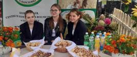 Trzy uśmiechnięte dziewczyny w ciemnych marynarkach siedzą przy stole wystawowym z poczęstunkiem. W tle widoczne logo szkoły rolniczej.