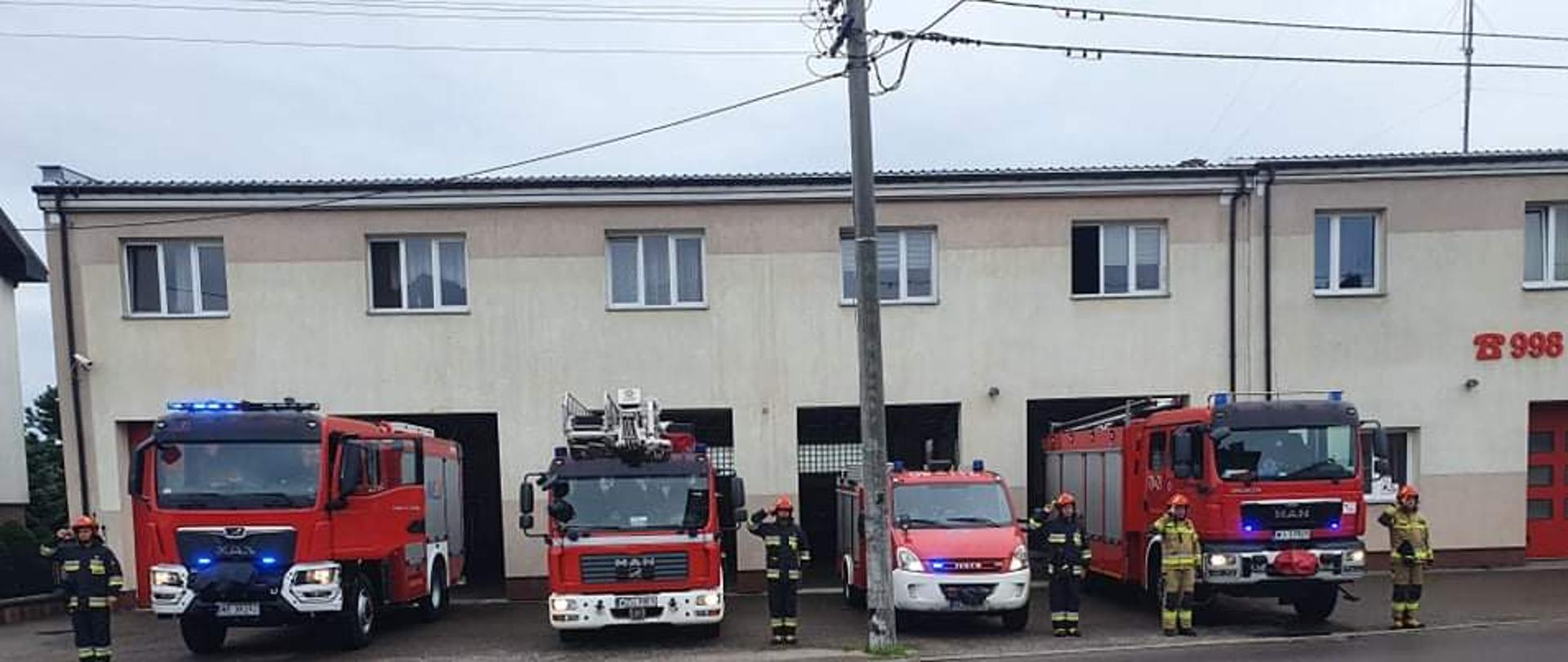 Przed budynkiem komendy stoją samochody ratowniczo gaśnicze z włączonymi sygnałami świetlno- dźwiękowymi. Przy samochodach stoją strażacy ubrani w stroje specjalne salutując.
