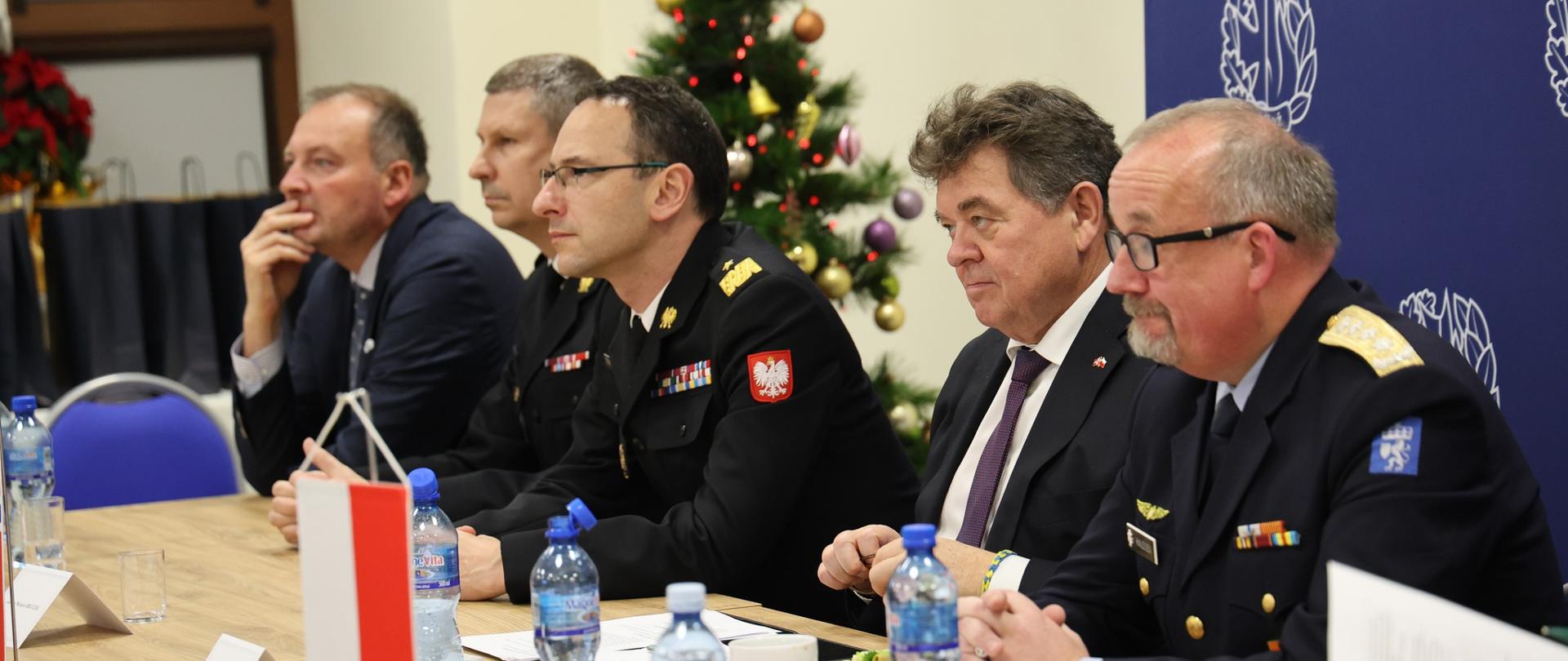 Pięciu mężczyzn siedzi przy stole konferencyjnym, w tym dwóch w mundurach PSP i jeden w mundurze zagranicznym; w tle choinka i ścianka z logo Akademii Pożarniczej.
