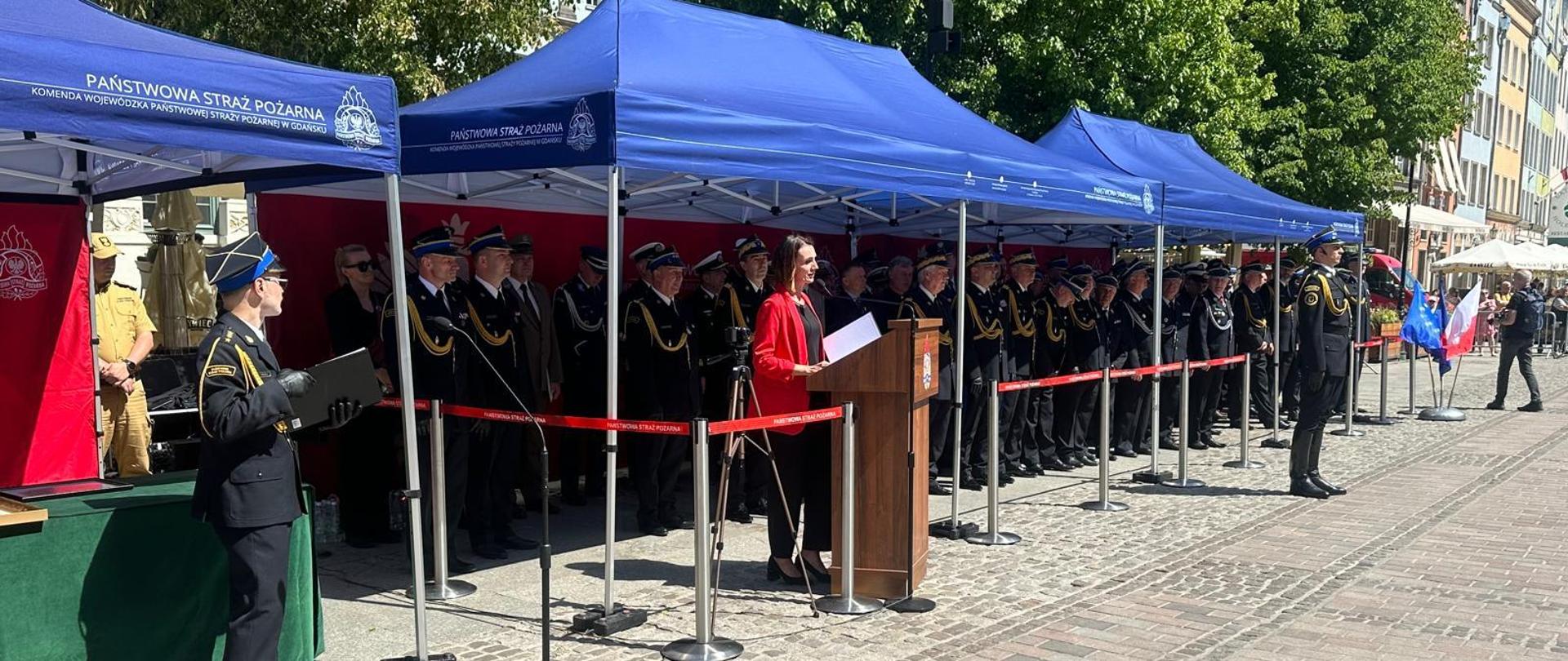 Grupa strażaków stoi pod namiotem na Długim Targu w Gdańsku, przed mikrofonem stoi wojewoda i przemawia