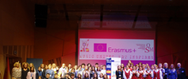 zdjęcie przedstawia uczestników programu Erasmus plus "Music Unites, Art Inspires". Uczestnicy ustawieni są na scenie sali koncertowej, w tle na ekranie wyświetlona jest grafika przedstawiająca na białym tle logo programu mobilności, logo programu Erasmus plus oraz logo Ogólnokształcącej Szkoły Muzycznej imienia Henryka Wieniawskiego w Łodzi 
