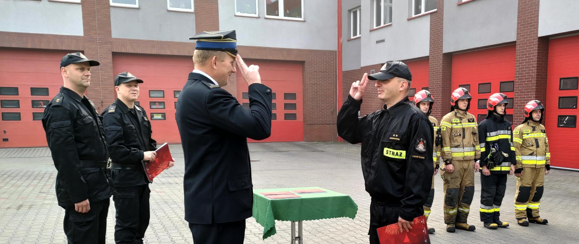 Zdjęcie przedstawia komendanta miejskiego i dowódcę zmiany oddających sobie honory na placu przed budyniem komendy. Obok stoi grupa strażaków. 