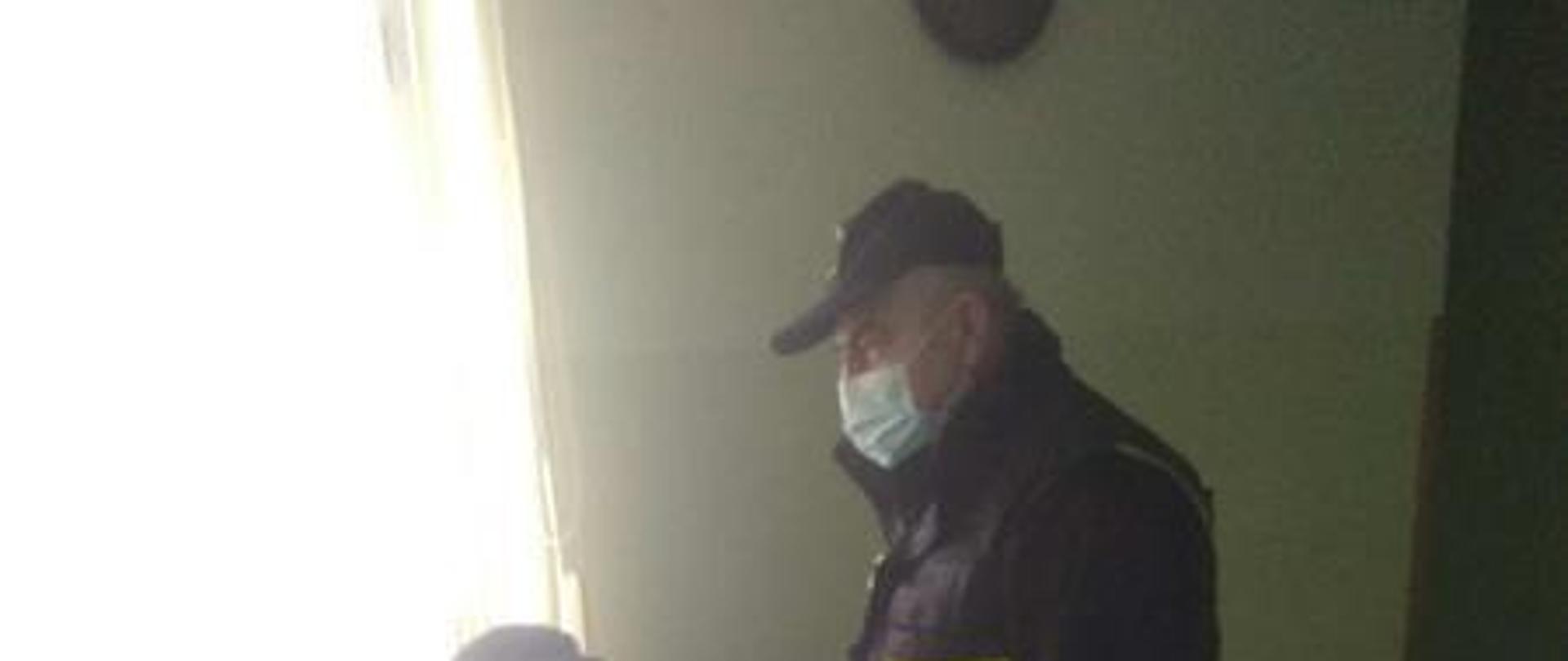 Osoba przygotowująca się do szczepienia siedzi na krzesełku, obok niej stoi strażak ubrany w rękawiczki oraz ma założoną maseczkę na twarz.