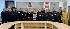 Grupa strażaków OSP oraz funkcjonariuszy PSP pozująca do pamiątkowego zdjęcia grupowego w sali szkoleniowej po pomyślnie zdanym egzaminie kończącym szkolenie kierujących działaniem ratowniczym. 