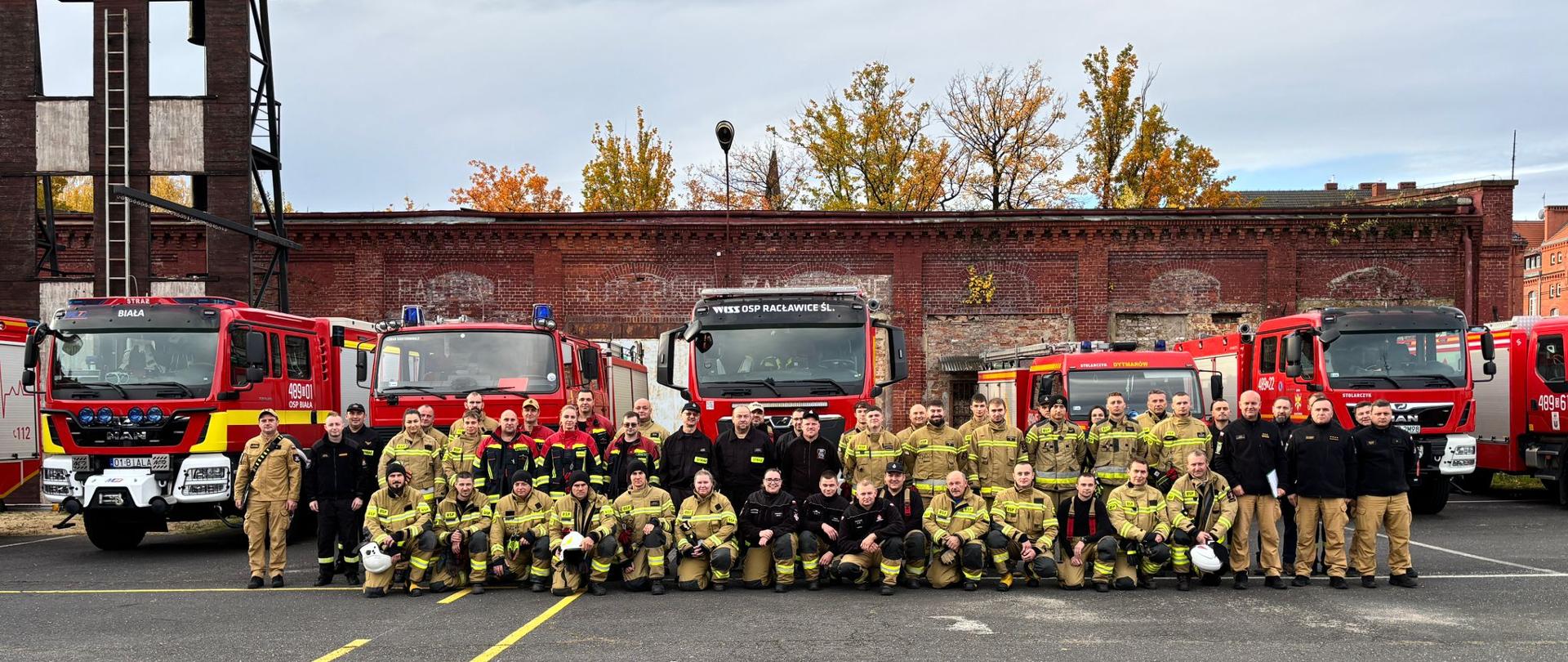 Zdjęcie przedstawia strażaków w strojach bojowych podczas wspólnej fotografii po warsztatach ratowniczych. W tle samochody pożarnicze stojące przed budynkiem