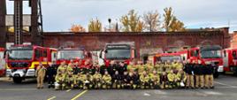 Zdjęcie przedstawia strażaków w strojach bojowych podczas wspólnej fotografii po warsztatach ratowniczych. W tle samochody pożarnicze stojące przed budynkiem