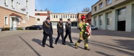 Kierownictwo Komendy Wojewódzkiej Państwowej Straży Pożarnej w Gdańsku w umundurowaniu wyjściowym idzie za strażakiem w umundurowaniu specjalnym, niosącym wieniec.