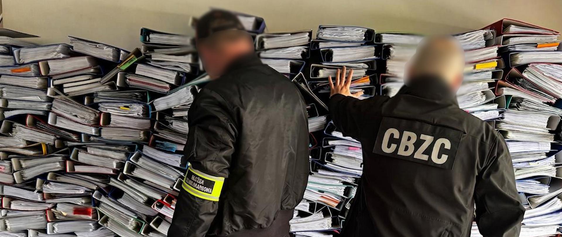 Dwie osoby stoją przed dużym stosem segregatorów i dokumentów ułożonych w nieregularne kolumny; jedna osoba dotyka dokumentów podczas kontroli.