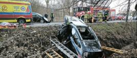 Czarny samochód osobowy leży w rowie po wypadku drogowym; strażacy prowadzą akcję ratunkową z użyciem drabin, na drodze stoją wóz strażacki i karetka pogotowia.