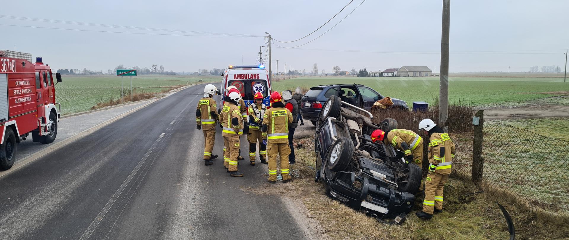 Zdjęcie przedstawia samochód osobowy leżący na dachu w przydrożnym rowie. Zderzak urwany leży przed samochodem. Na zdjęciu widać również strażaków oraz samochód strażacki i karetkę pogotowia ratunkowego. 