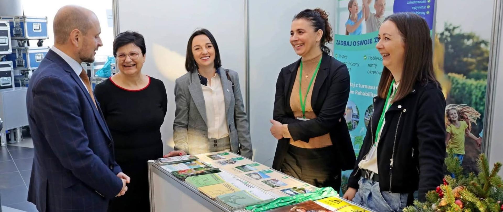 Mężczyzna i cztery kobiety stoją przy stoisku prewencyjnym Kasy – na stoliku wyłożone są materiały prewencyjne (ulotki). Za nimi znajduje się roll-up kasy.