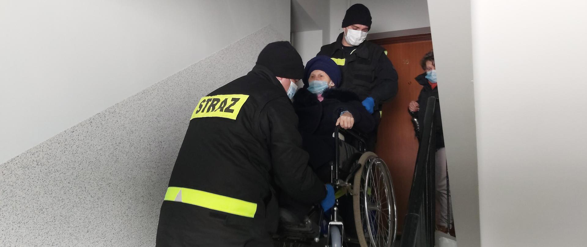 Dwóch strażaków znosi osobę na wózku inwalidzkim po schodach. Wszyscy w maskach na twarzy