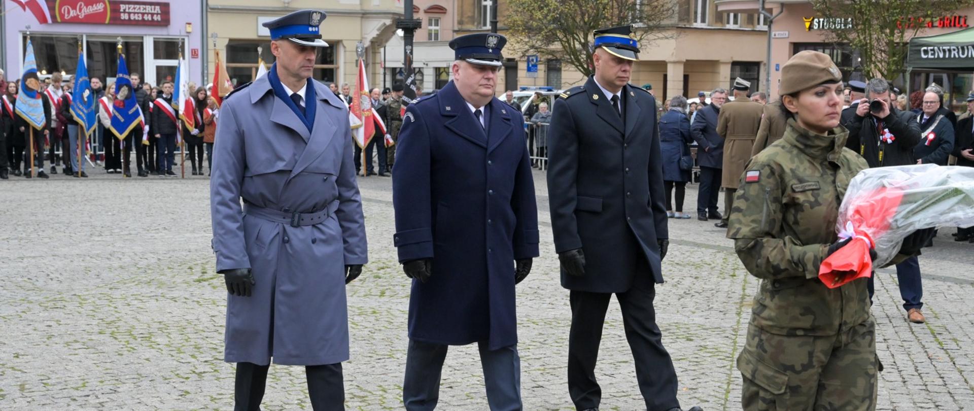 Zdjęcie przedstawia wybrukowany plac, na którym zgromadzeni są ludzie. Na pierwszym planie maszeruje 3-osobowa delegacja mężczyzn w mundurach, którą poprzedza kobieta w mundurze moro niosą ca wiązankę kwiatów.