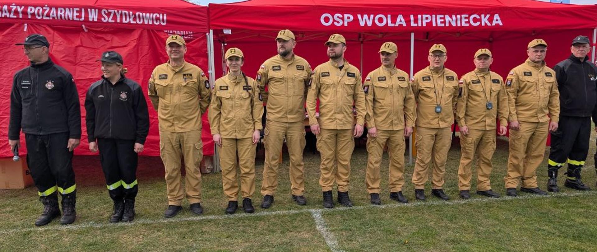 Zdjęcie przedstawia grupę funkcjonariuszy oraz druhów podczas wydarzenia plenerowego, prawdopodobnie zawodów sportowo-pożarniczych.
• Uczestnicy: W jednym szeregu stoi jedenaście osób. Większość ubrana jest w piaskowe mundury służbowe z czapkami z daszkiem. Dwie osoby po lewej oraz jedna po prawej noszą czarne mundury ćwiczebne.
• Tło: Za plecami zebranych rozstawione są duże, czerwone namioty promocyjne. Na jednym z nich widnieje biały napis „OSP WOLA LIPIENIECKA”, a na sąsiednim fragment napisu dotyczący straży w Szydłowcu.
• Otoczenie: Akcja dzieje się na trawiastym boisku lub placu, w słoneczny dzień. Na trawie widać namalowane białe linie wyznaczające pole startowe lub sektory.
