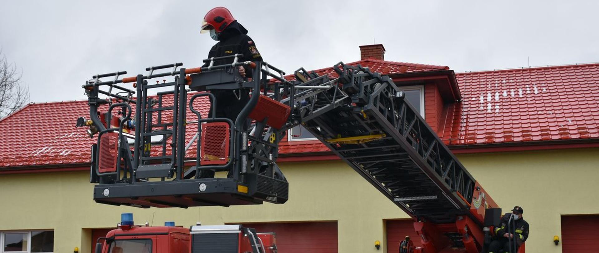 Na zdjęciu widzimy wysuniętą drabinę mechaniczną na samochodzie. Jeden ze strażaków znajduje się w koszu drabiny, natomiast drugi strażak siedzi przy pulpicie sterowniczym drabiny. Strażacy ubrani są w ubrania specjalne. Strażak znajdujący się w koszu ma również hełm na głowie. Samochód z drabina mechaniczna stoi na tle budynku strażnicy.