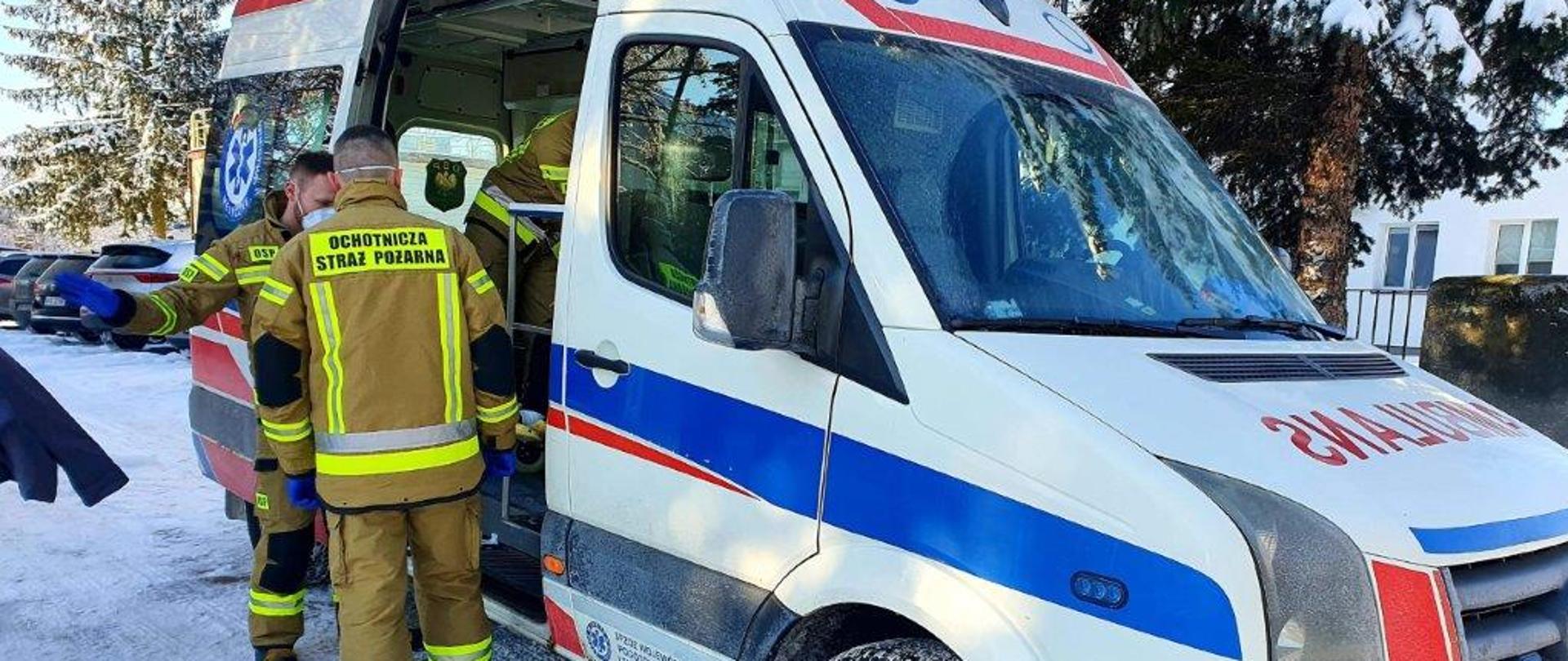 Strażacy przy ambulansie, pomagają wysiąść starszej osobie.