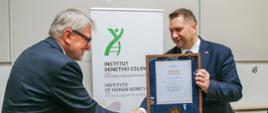 Minister Czarnek ściska rękę siwego mężczyzny w garniturze, w drugiej ręce trzyma dokument w ramkach, za nimi przed białą ścianą baner z napisem Instytut Genetyki Człowieka.