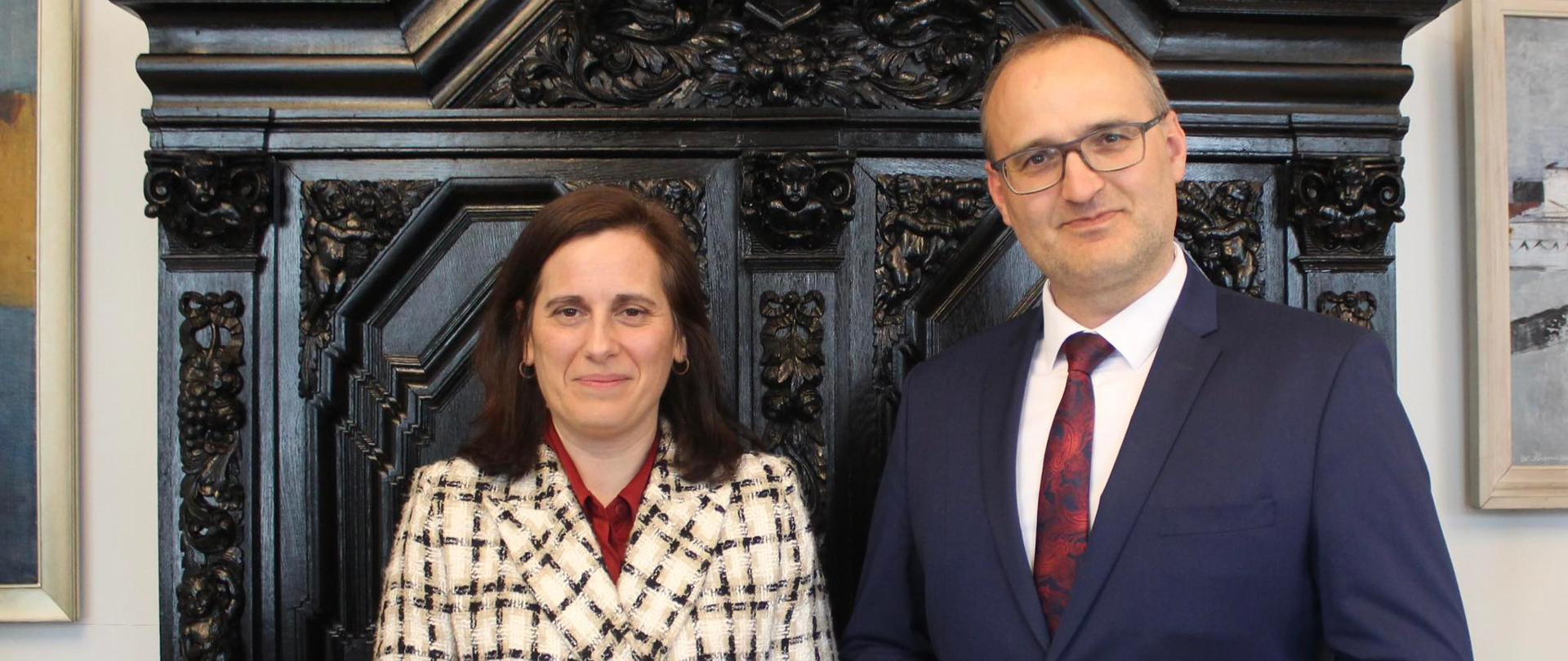 Zdjęcie przedstawia dwoje ludzi - kobietę i mężczyznę - stojących obok siebie w oficjalnych strojach na tle ciemnego mebla.
