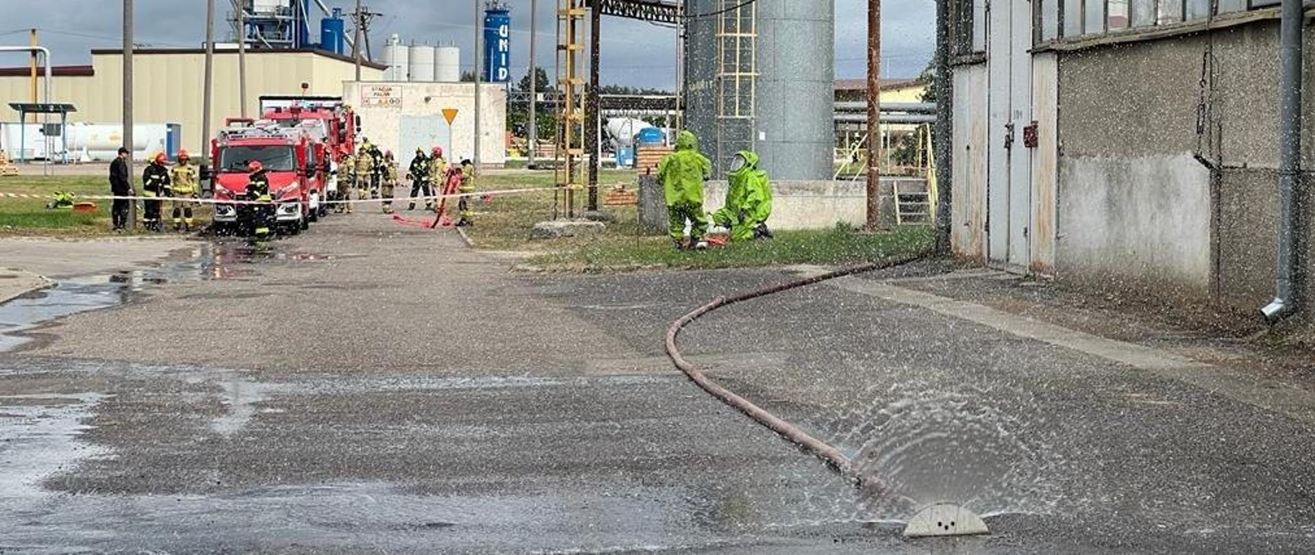 Rozstawiona kurtyna wodna z której płynie woda. W tle pracują strażacy w ubraniach gazoszczelnych. Z lewej stoją samochody straży pożarnej oraz strażacy.