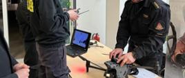 Zdjęcie wykonane na korytarzu budynku Leśnego Ośrodka Edukacyjnego w Jedlni Letnisko. Na zdjęciu widać ustawione biurko. Na biurku znajduje się laptop, urządzenie przeznaczone do sterowania oraz bezzałogowy statek powietrzny – dron. Po prawej stronie biurka znajduje się funkcjonariusz Państwowej Straży Pożarnej. Strażak posiada uprawnienia operatora drona. Ubrany jest w ubranie dowódczo – sztabowe w kolorze czarnym. Po lewej stronie widać innych funkcjonariuszy Państwowej Straży Pożarnej, którzy przyglądają się prezentowanemu sprzętowi. Strażacy ubrani są w ubrania koszarowe w kolorze czarnym z odblaskowymi elementami. 