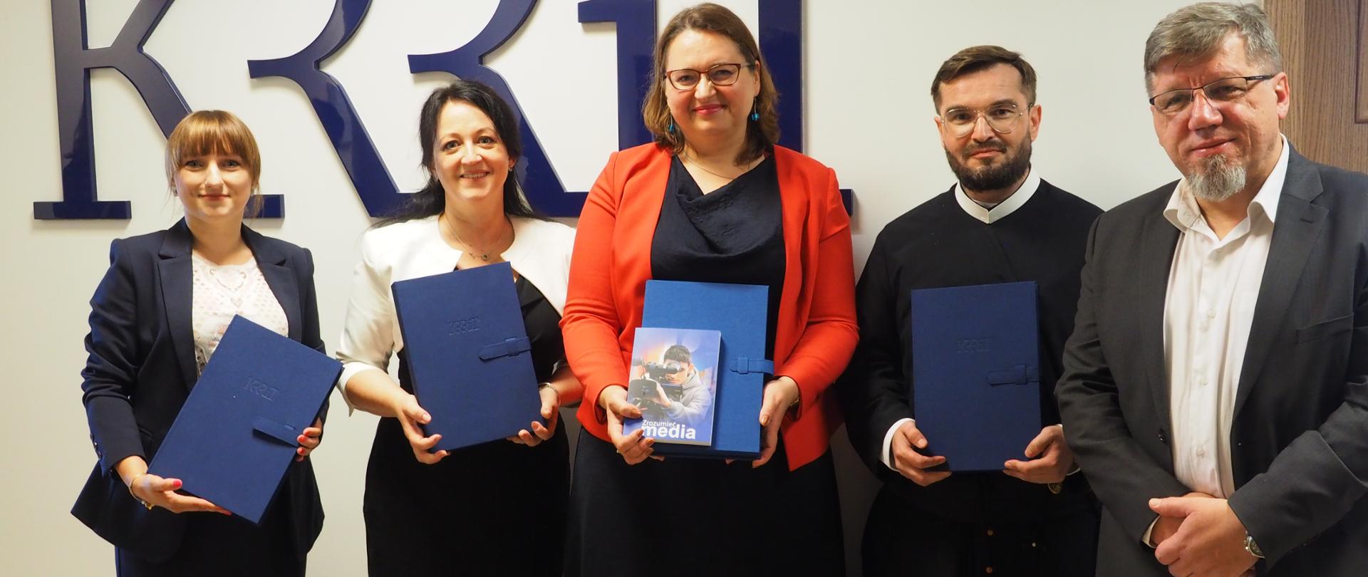 Laureaci Nagrody im. dr. Pawła Stępki z Przewodniczącym KRRiT stoją z dyplomami pod niebieskim logo KRRiT
