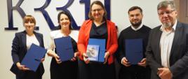 Laureaci Nagrody im. dr. Pawła Stępki z Przewodniczącym KRRiT stoją z dyplomami pod niebieskim logo KRRiT