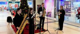 Kwartet saksofonowy gra przy udekorowanej choince w pasażu galerii handlowej.