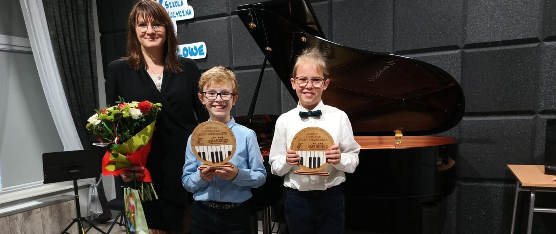 Dwóch uczniów stojących, trzymających statuetki laureatów wraz z nauczycielką trzymającą bukiet kwiatów na tle czarnej ściany. Z tyłu widoczny jest fortepian oraz pulpit po lewej stronie. 