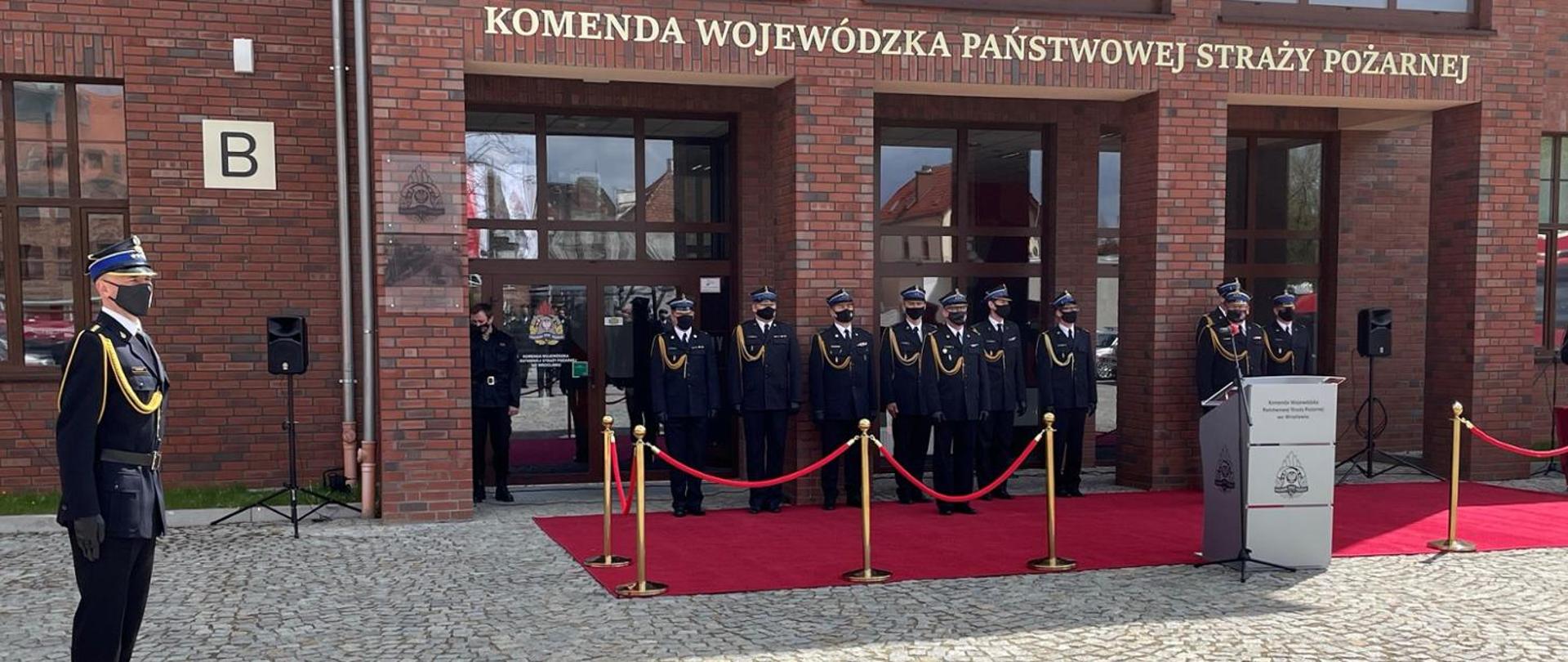 Zdjęcie przedstawia trybunę honorową podczas uroczystości zakończenia szkolenia podstawowego w zawodzie strażak. W tle budynek Komendy Wojewódzkiej Państwowej Straży Pożarnej we Wrocławiu. Po lewej stronie zdjęcia widoczny dowódca uroczystości. Wszyscy strażacy ubrani są w ubrania galowe - mundur wyjściowy.