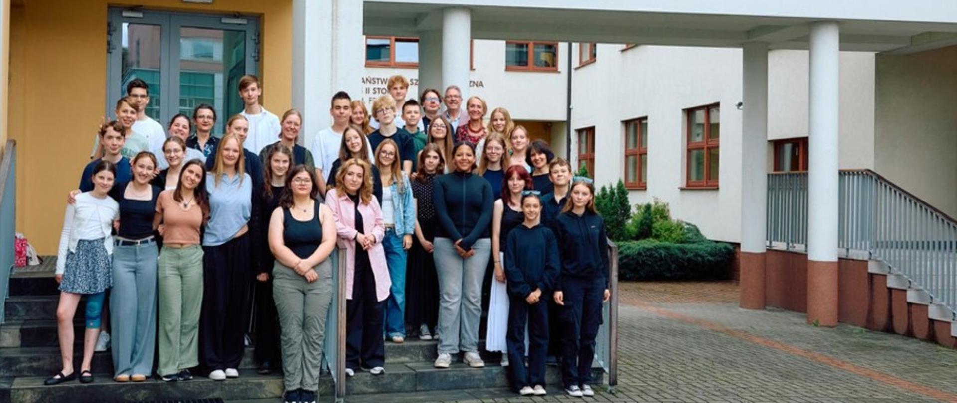 Na zdjęciu widoczna jest duża grupa młodzieży i nauczycieli, którzy stoją wspólnie przed wejściem do budynku szkoły muzycznej. Budynek jest jasny, nowoczesny, z dużymi oknami i przeszklonymi elementami.Na ścianie po prawej stronie widnieje napis:
Państwowa Szkoła Muzyczna I i II stopnia im. Fryderyka Chopina w Nowym Targu, obok którego znajduje się ozdobne logo. Po lewej stronie zdjęcia, nad wejściem, znajdują się logotypy: kolorowe logo Małopolski, logo Polsko-Niemieckiej Współpracy Młodzieży (Deutsch-Polnisches Jugendwerk). Grupa, licząca około 35 osób, stoi na schodach i przed wejściem, ustawiona w kilku rzędach. Widać uśmiechnięte twarze, część osób obejmuje się ramionami, co podkreśla przyjazną i koleżeńską atmosferę. Zdjęcie ma charakter pamiątkowy i oficjalny, prawdopodobnie wykonane zostało przy okazji projektu wymiany młodzieży lub wydarzenia artystyczno-edukacyjnego.