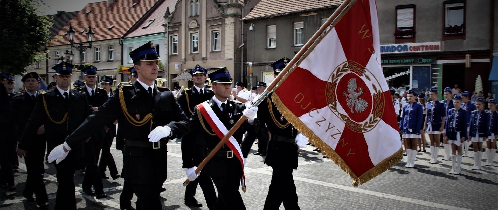 Zdjęcie z uroczystości nadania sztandaru dla Komendy Powiatowej Państwowej Straży Pożarnej w Ostrzeszowie w 2013 roku. Na pierwszym tle poczet sztandarowy w trakcie prezentowania sztandaru w marszu defiladowym. Na drugim planie pododdział funkcjonariuszy Komendy Powiatowej.