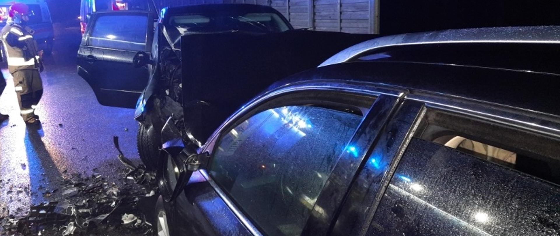 Na zdjęciu widać dwa samochody osobowe które zderzyły się czołowo po lewej stronie stoi strażak w umundurowaniu bojowym w tle dwie karetki pogotowia ratunkowego i ekrany dźwiękochłonne