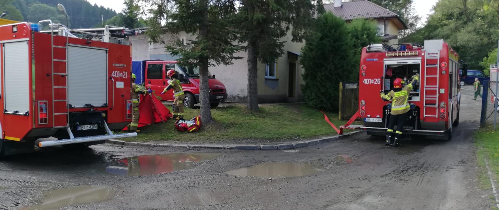Na zdjęciu widać dwa pojazdy pożarnicze oraz trzech strażaków w ubraniach specjalnych. Jeden z nich obsługuje autopompę, dwóch kolejnych rozkłada parawan, obok nich rozłożona jest torba R1. Z pojazdu którego strażak obsługuje autopompę rozwinięty jest wąż pożarniczy. Za samochodami stoi budynek MPGK w Ustrzykach Dolnych oraz stoi pojazd należący do MPGK. 