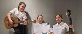 Trzy uśmiechnięte dziewczynki na tle szarej ściany prezentują swoje nagrody muzyczne zdobyte podczas konkursu gitarowego. Uczennice ubrane są w galowe stroje – białe bluzki i ciemne spodnie. Dziewczynka po lewej stronie stoi, trzymając opartą o kolano gitarę klasyczną. Dziewczynka w środku kuca, trzymając w rękach dwa dyplomy (w tym dyplom za III Miejsce dla Zofii Nowak). Dziewczynka po prawej stronie klęczy, trzyma przed sobą dwa dyplomy, a obok niej stoi oparta o podłogę duża gitara klasyczna.