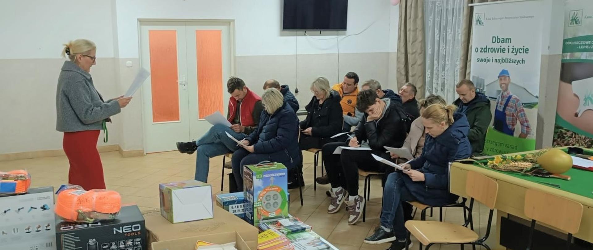 Uczestnicy siedzą na krzesłach i rozwiązują test wiedzy. Za nimi znajdują się roll-upy KRUS. Przed nimi stoi Małgorzata Pyra, która nadzoruje przebieg testu.