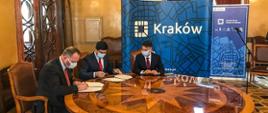 Trzech mężczyzn siedzi przy okrągłym stole i podpisują dokumenty. Umowa dotyczy budowy linii tramwajowej do Mistrzejowic. W tle ścianka z napisem Kraków.