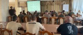 Grupa mężczyzn siedzi plecami do kamery w trzech rzędach ławek i patrzy na prezentację wyświetlaną na ekranie, obok którego stoi stół, przy którym siedzą szkoleniowcy; z drugiej trony stołu jest tablica flipchart z umieszczonym na niej dokumencie