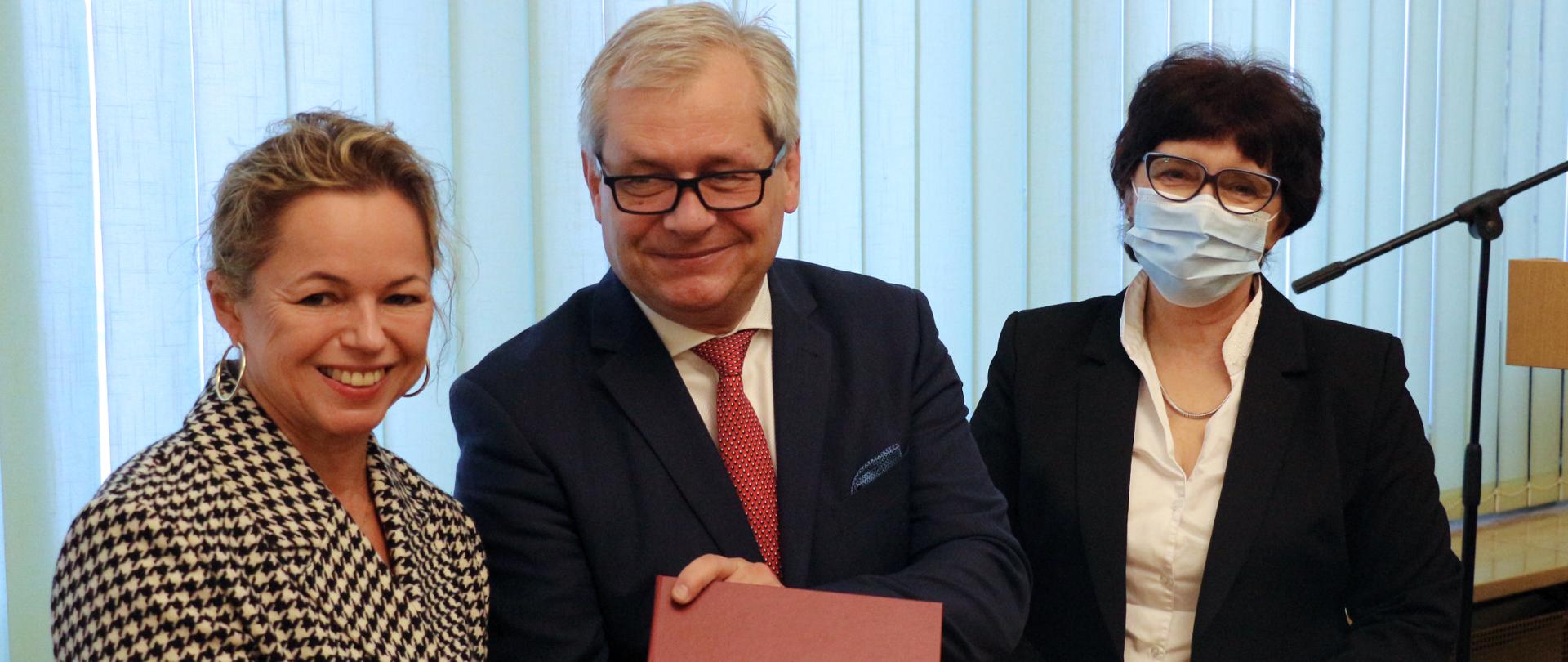 Na zdjęciu trzy osoby, dwie kobiety oraz wojewoda opolski. Bliski kadr. Cała trójka pozuje do zdjęcia. Kobieta po lewej stronie kadru trzyma w dłoniach czerwoną teczkę ze złotym orzełkiem i napisem Wojewoda Opolski. Kobieta ubrana jest w kraciastą marynarkę, ma blond włosy, jest młoda i spogląda do obiektywu aparatu. Obok niej stoi wojewoda opolski, który spogląda na kobietę, uśmiecha się. Na twarzy ma okulary. Po prawej stronie kadru stoi ubrana w elegancki, ciemny strój przedstawicielka resortu rodziny i polityki społecznej. Kobieta ma ciemne, krótkie włosy, okulary korekcyjne. Na szyi ma naszyjnik z pereł. Pozując do zdjęcia, dłonie trzyma w uścisku. W tle okno oraz rolety.