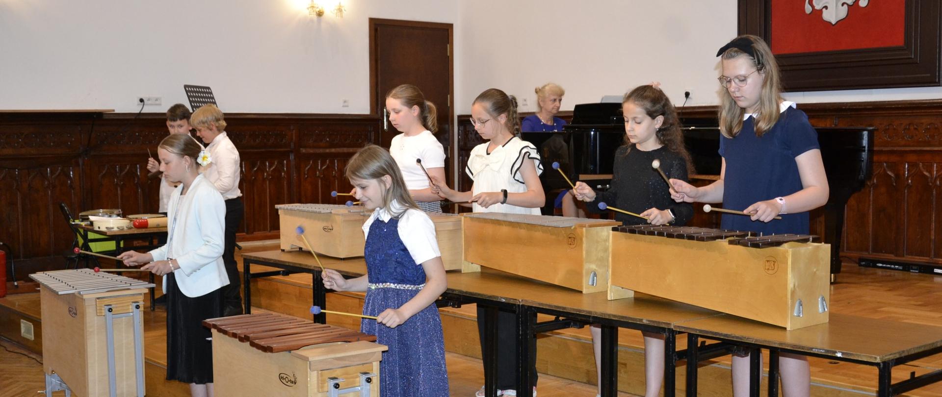 Uczennice grają na ksylofonach i metalofonie podczas koncertu z okazji Dnia Matki w Sali Królewskiej, w tle nauczycielka przy fortepianie i herb Polski.