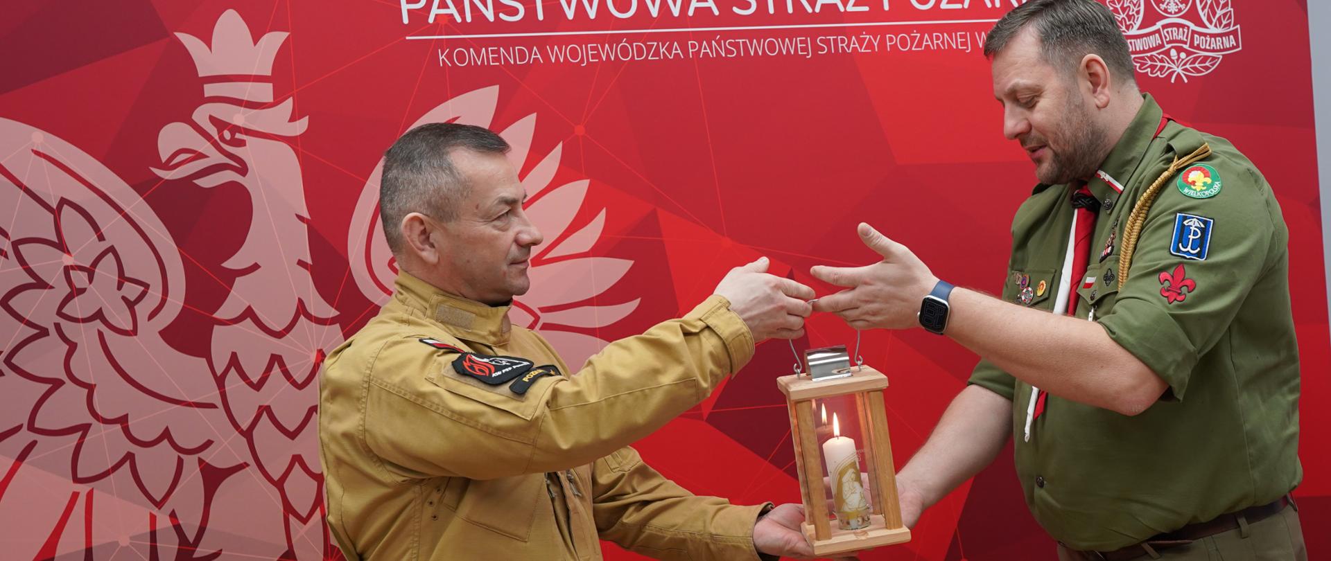 Dwóch mężczyzn w mundurach służbowych strażak i harcerz, przekazuje sobie zapaloną świecę umieszczoną w drewnianej latarni. Stoją na tle czerwonej ścianki z napisem „Państwowa Straż Pożarna” oraz godłem. Scena wygląda na oficjalne, uroczyste przekazanie symbolicznego ognia.