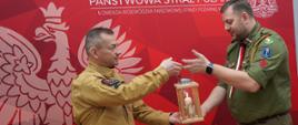 Dwóch mężczyzn w mundurach służbowych strażak i harcerz, przekazuje sobie zapaloną świecę umieszczoną w drewnianej latarni. Stoją na tle czerwonej ścianki z napisem „Państwowa Straż Pożarna” oraz godłem. Scena wygląda na oficjalne, uroczyste przekazanie symbolicznego ognia.