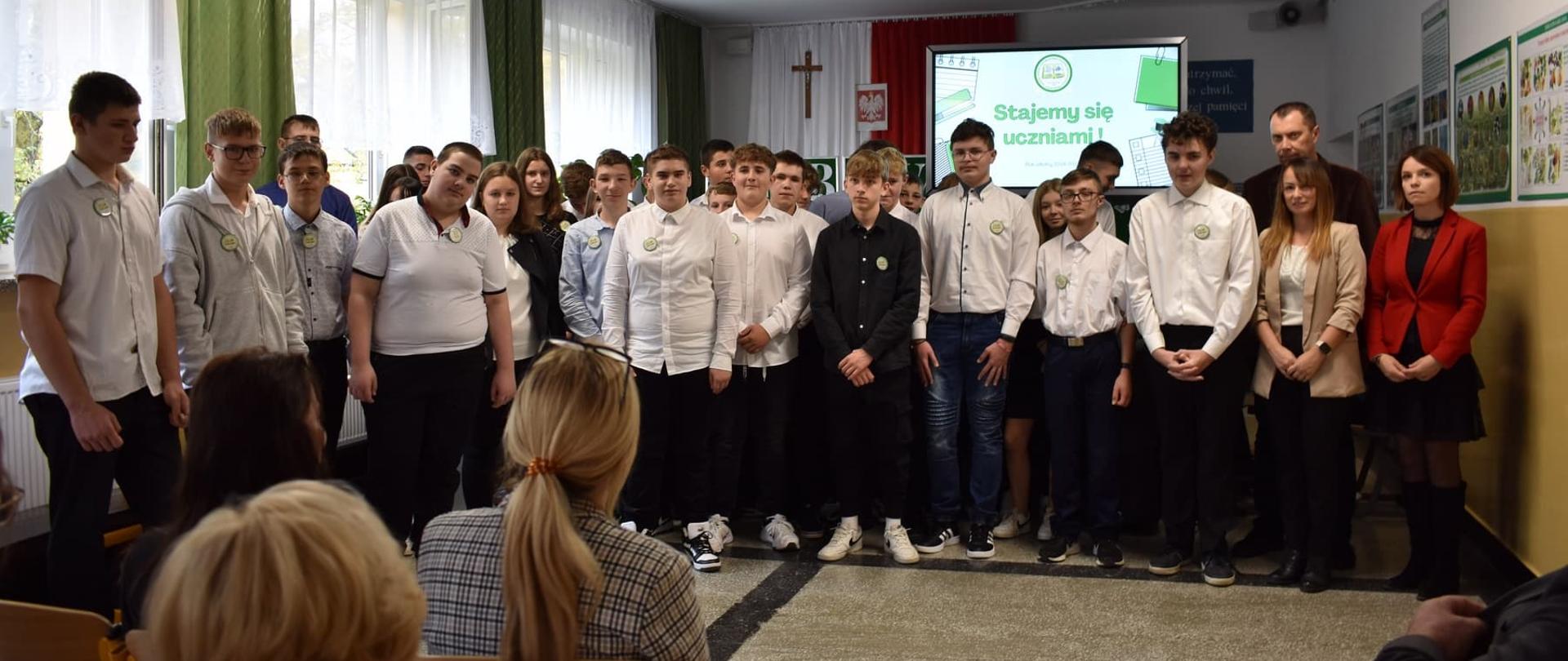 Zbiorowe zdjęcie uczniów i nauczycieli stojących przed ekranem z napisem „Stajemy się uczniami!”.