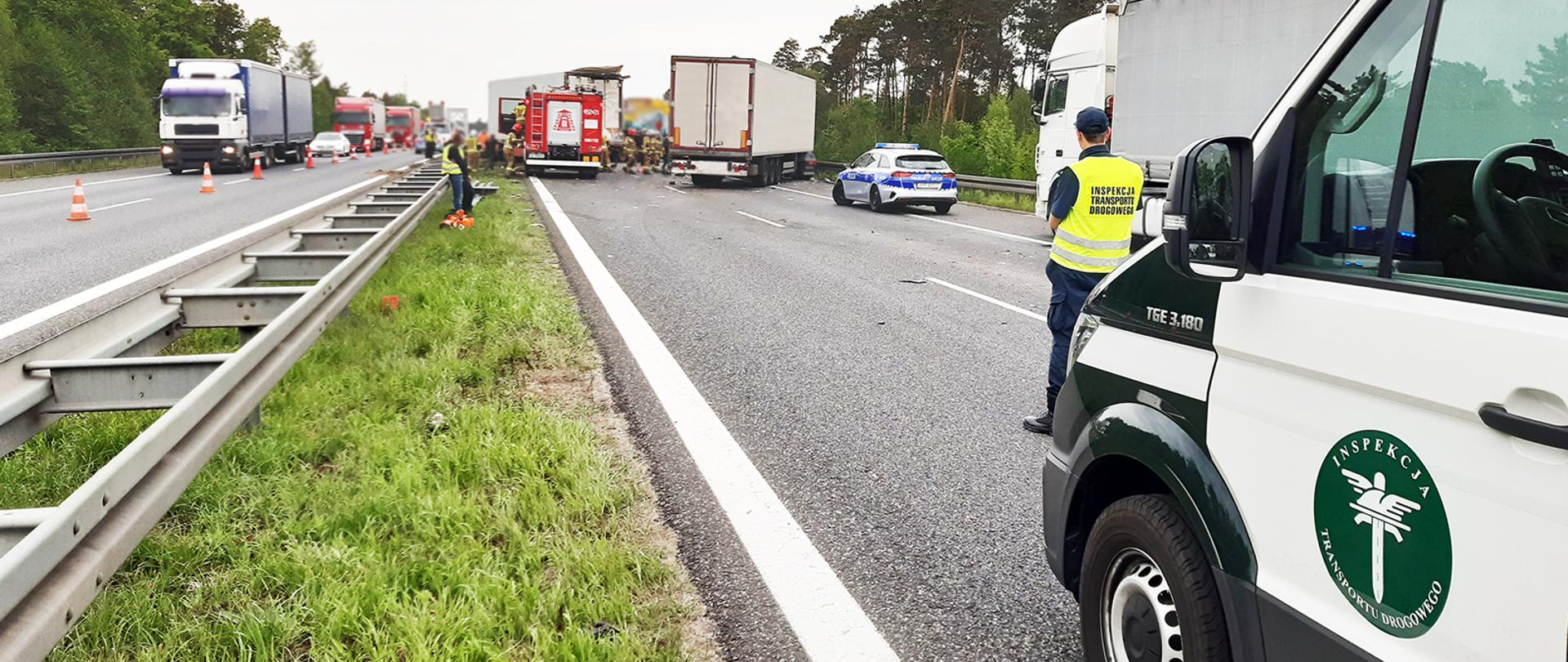 Inspektorzy śląskiej ITD na miejscu tragicznego zdarzenia. Na pierwszym planie inspekcyjny furgon i inspektor ITD. W centrum miejsce tragicznego zdarzenia - rozbite pojazdy i służby (straż pożarna i policja). Po lewej ruch pojazdów.