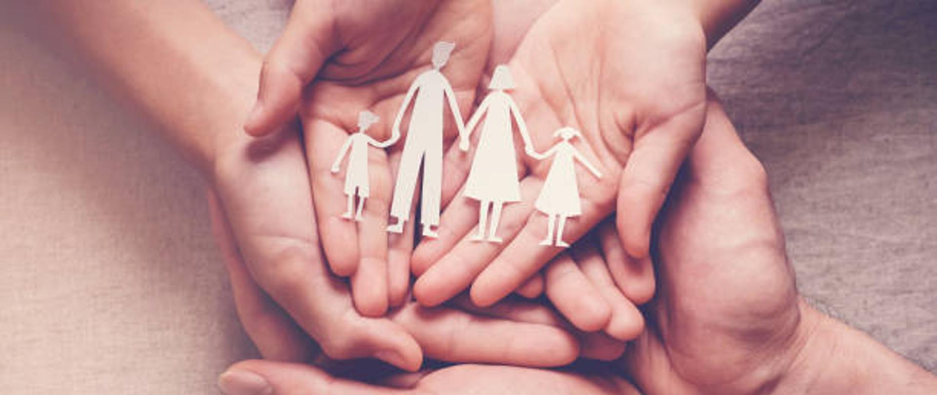 ułożone na sobie otwarte dłonie trzech osób - od największej do najmniejszej, a na nich wycięte z papieru figurki czteroosobowej rodziny